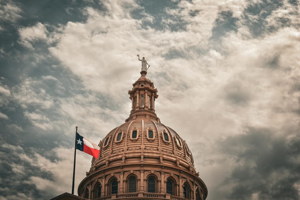 The dome of the Texas State Capitol with a Texas flag under a cloudy sky.