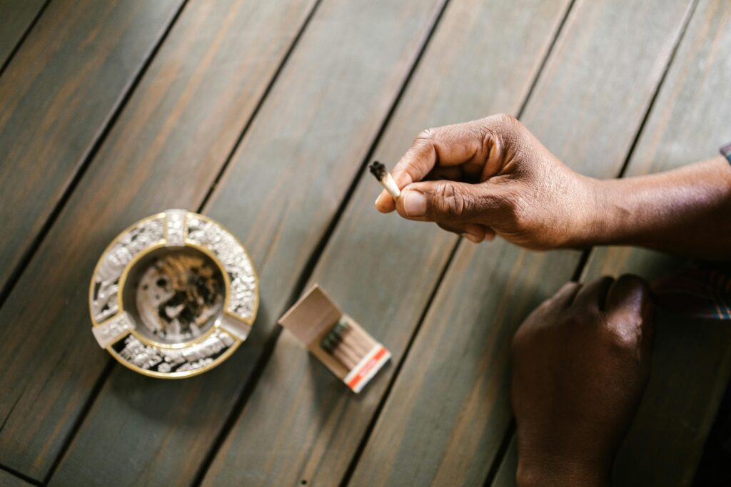 A close-up shot of a person holding a marijuana joint above a wooden table with ashtray and matches.